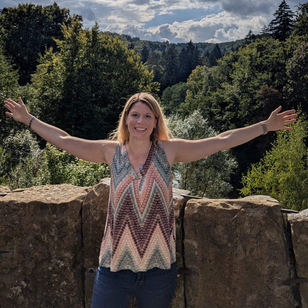 Yvonne Weilandt steht lachend mit ausgebreiteten Armen vor Wald und Himmel